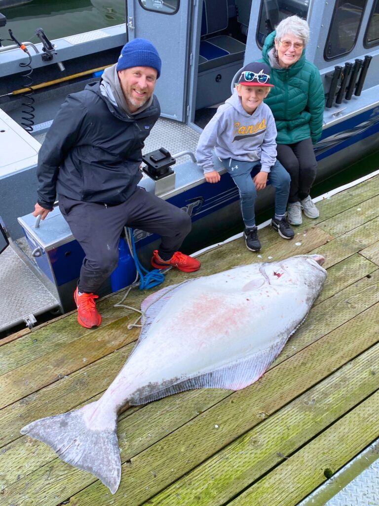 A family with a young boy posing with a large halibut at the dock beside their rental boat.
