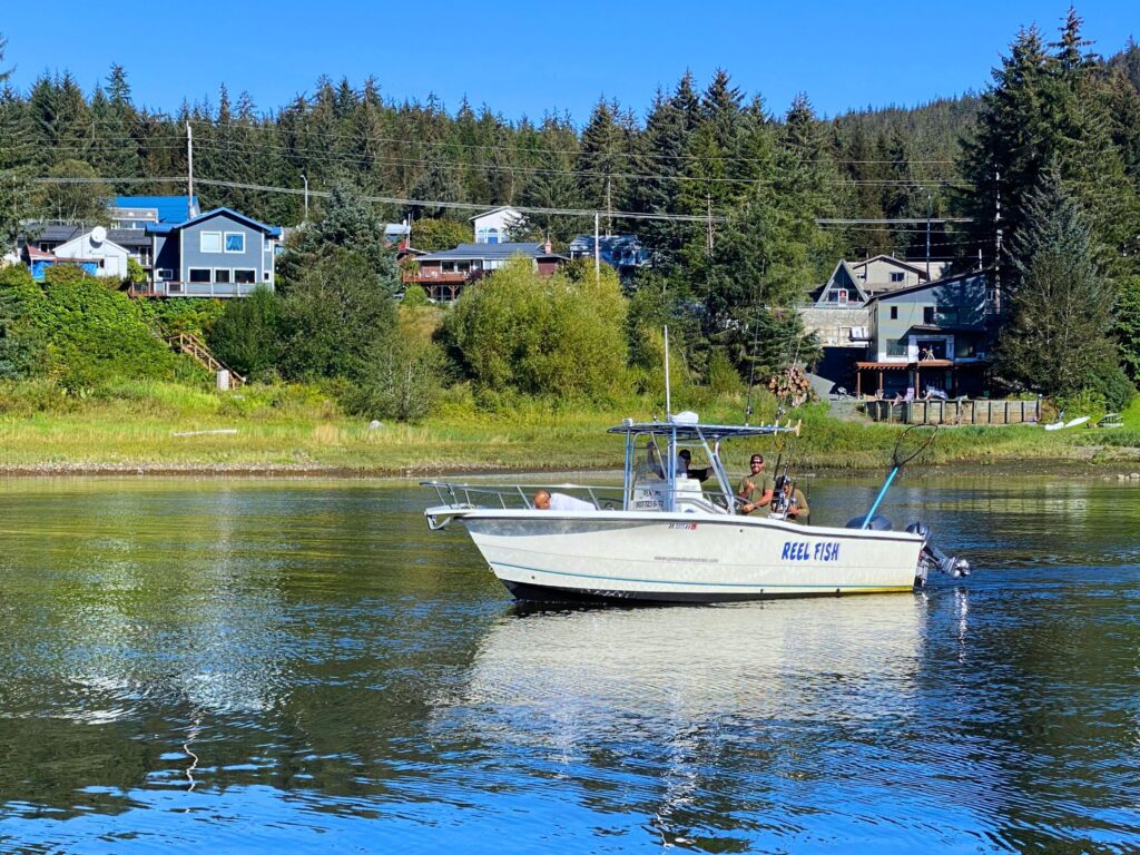Two anglers aboard the Reel Fish boat fishing on calm water near a forested Southeast Alaska shoreline.