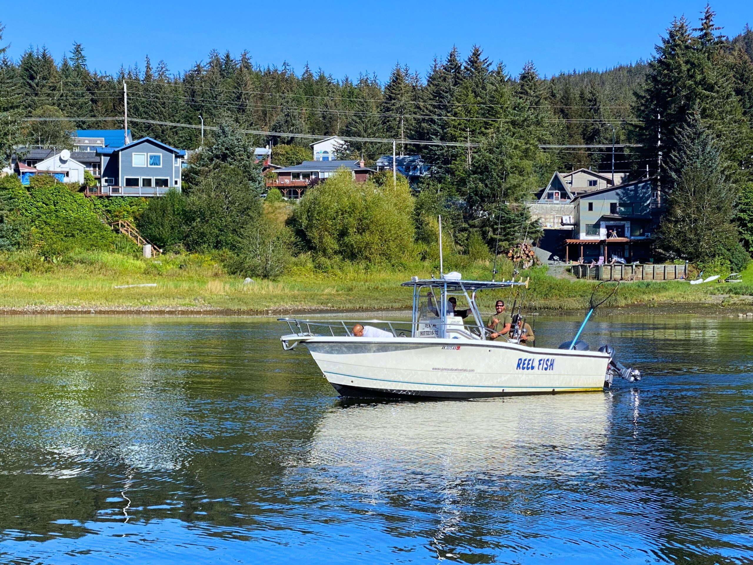 Two anglers aboard the Reel Fish boat fishing on calm water near a forested Southeast Alaska shoreline.